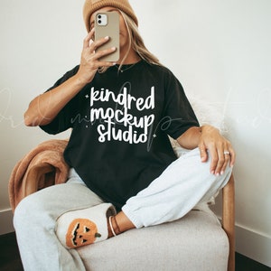 Op de afbeelding: Een vrouw draagt een zwart T-shirt met de tekst "kindred mockup studio" in witte letters. Ze zit op een stoel met gekruiste benen en draagt een paar pantoffels met een pompoenthema.
