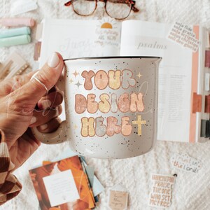 May include: A speckled white ceramic mug with the text "YOUR DESIGN HERE" in retro-style lettering. The mug is held in a hand, with a Bible and other stationery items in the background.