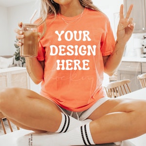 May include: A woman wearing a coral colored t-shirt with the text "YOUR DESIGN HERE" in white. She is sitting on a white surface with her legs crossed and holding a glass jar with a straw.