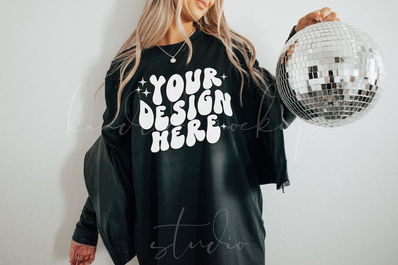 May include: Black long-sleeve shirt with the text "YOUR DESIGN HERE" in white, with star accents. A silver disco ball is held in the right hand. The shirt is paired with a black jacket.