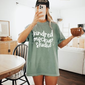 May include: A woman wearing a sage green t-shirt with the text "kindred mockup studio" printed in white. She is holding a brown mug in her right hand and a cell phone in her left hand.