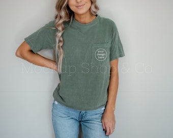 Op de afbeelding: Een vrouw draagt een groen T-shirt met een zakje waarop "your design here" staat. Ze draagt een blauwe jeans en heeft haar hand op haar heup.