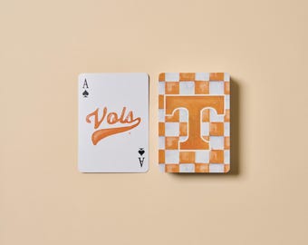 May include: Two playing cards on a tan surface. One card is the Ace of Spades with the word "Vols" in orange script. The other card has a checkerboard pattern in orange and white with a large orange "T".