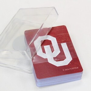 Puede incluir: Un mazo de cartas en una caja de pl&aacute;stico transparente. Las cartas son de color granate con un logotipo "OU" blanco y el texto "&copy;2024 FOSTER". La caja est&aacute; abierta, mostrando las cartas.