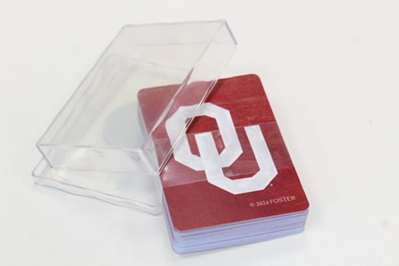 Puede incluir: Un mazo de cartas en una caja de pl&aacute;stico transparente. Las cartas son de color granate con un logotipo "OU" blanco y el texto "&copy;2024 FOSTER". La caja est&aacute; abierta, mostrando las cartas.