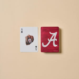 May include: A playing card showing the Ace of Spades with a fist wearing a ring, and a red card with a white "A" logo. The cards are on a beige background. The deck of cards has a sports theme.