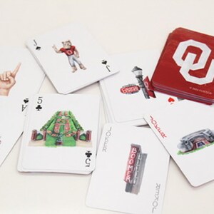 Puede incluir: Una baraja de cartas con varias ilustraciones. Las cartas presentan im&aacute;genes de un estadio, un gesto con la mano, una mascota y el texto "BOOMER". Los dorsos de las cartas son rojos con un logotipo blanco.