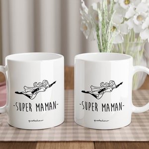 May include: Two white ceramic mugs with black handles. Each mug features a black and white illustration of a woman flying with the text "SUPER MAMAN" printed below. The mugs are sitting on a checkered table cloth.
