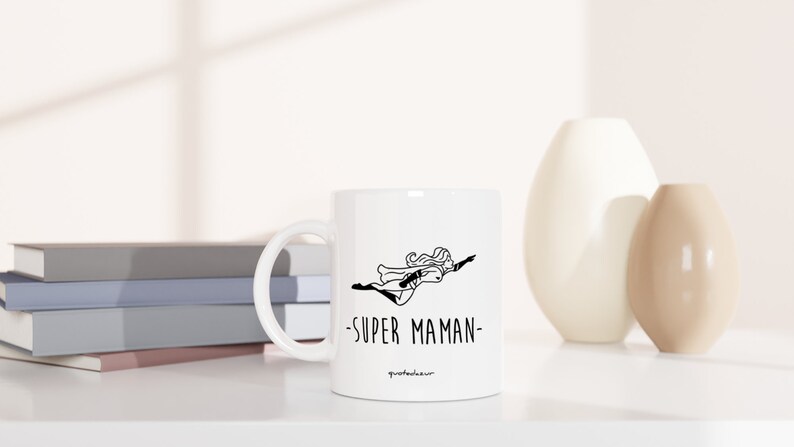 May include: White ceramic mug with a black line drawing of a woman flying with the text "SUPER MAMAN" on the front.