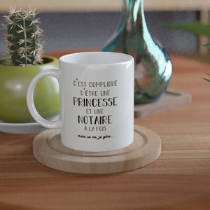 May include: White ceramic coffee mug with a black inscription that reads "C'est compliqu&eacute; d'&ecirc;tre une princesse et une notaire &agrave; la fois mais &ccedil;a va, je g&egrave;re..." The mug is sitting on a wooden coaster on a wooden table.