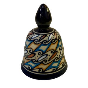 May include: A ceramic bell with a black top and base, and a patterned body. The body features a design of blue, orange, and white shapes on a beige background. The bell is a decorative item.