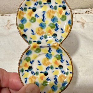 May include: A small, double-dish ceramic condiment server. The white surface is decorated with a floral pattern in yellow, blue, green, and black. The rim is trimmed with gold. The server is held by a hand.