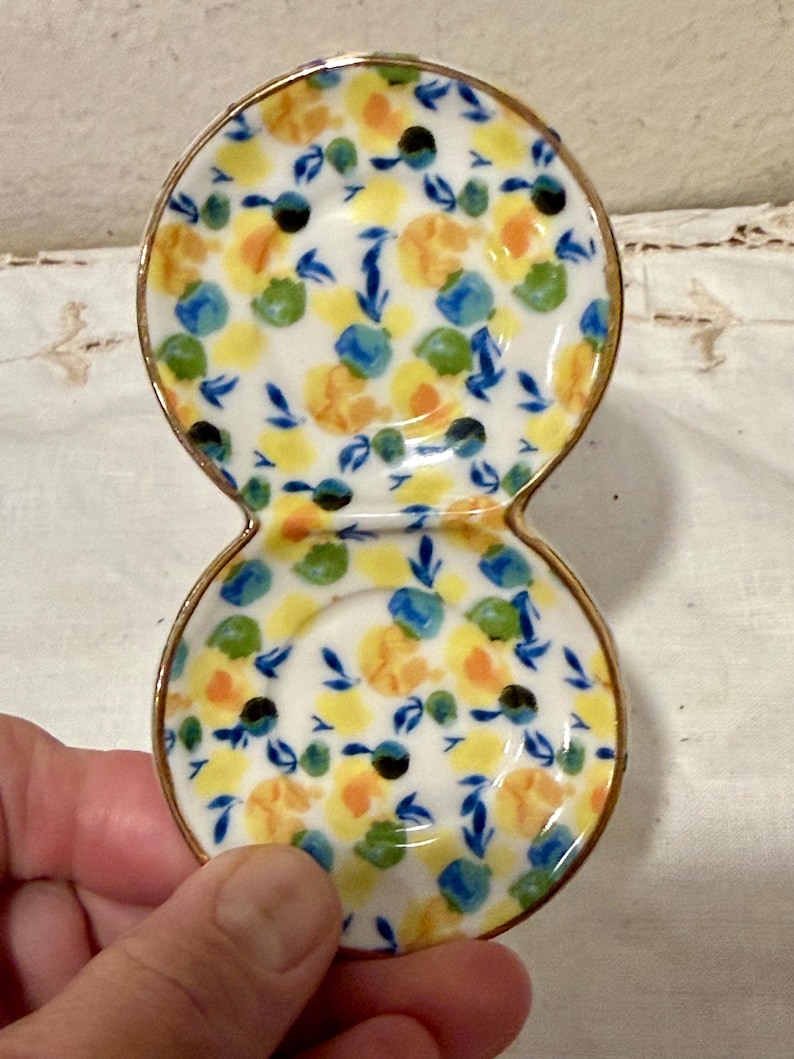 May include: A small, double-dish ceramic condiment server. The white surface is decorated with a floral pattern in yellow, blue, green, and black. The rim is trimmed with gold. The server is held by a hand.