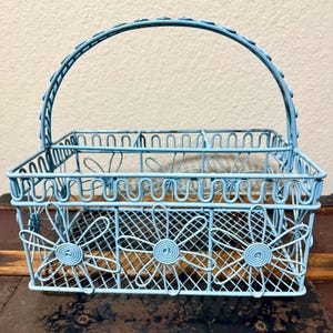 May include: A light blue wire basket with a handle. The basket has a rectangular shape with floral designs on the sides and a handle that arches over the top. The basket is made of metal.