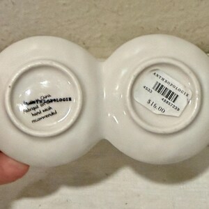 May include: A white ceramic Anthropologie dish with two connected circular compartments. The bottom of the dish has text that reads "Made in China, Fabriqu&eacute; en Chine, hand wash recommended."
