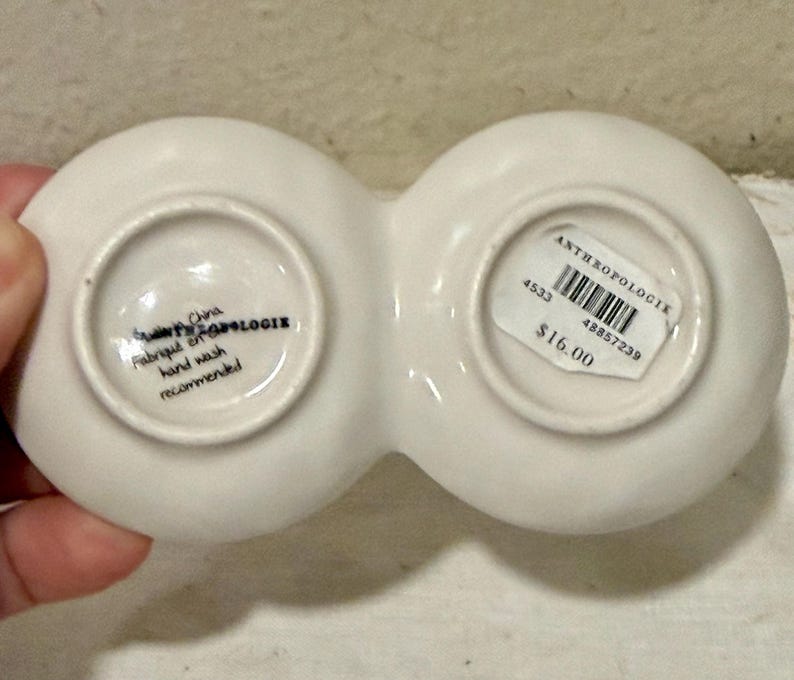 May include: A white ceramic Anthropologie dish with two connected circular compartments. The bottom of the dish has text that reads "Made in China, Fabriqu&eacute; en Chine, hand wash recommended."