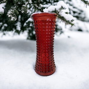 May include: A red glass vase with a textured surface sits in the snow. The vase is tall and narrow with a rounded bottom.