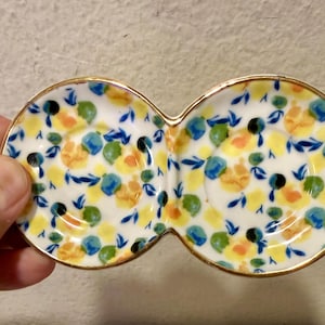 May include: A white ceramic serving dish with a double-bowl design. The dish is decorated with a floral pattern in yellow, blue, green, and black. The rim is trimmed with gold.