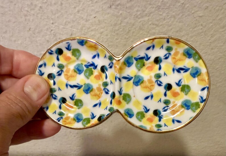 May include: A white ceramic serving dish with a double-bowl design. The dish is decorated with a floral pattern in yellow, blue, green, and black. The rim is trimmed with gold.