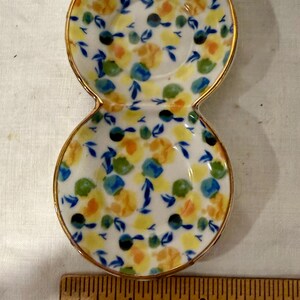 May include: A small, double-dish ceramic plate with a gold rim. The plate is decorated with a floral pattern in yellow, blue, green, and black on a white background. A wooden ruler with inch markings is in the foreground.