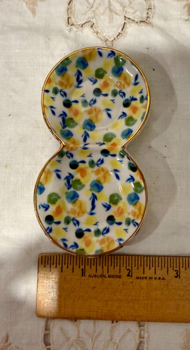 May include: A small, double-dish ceramic plate with a gold rim. The plate is decorated with a floral pattern in yellow, blue, green, and black on a white background. A wooden ruler with inch markings is in the foreground.