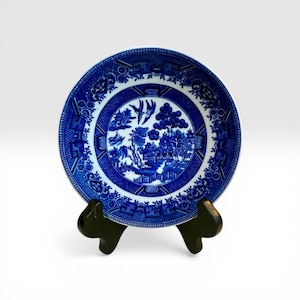 May include: A blue and white ceramic plate with a Willow pattern design. The plate features a detailed landscape scene with pagodas, trees, and birds. The plate is displayed on a dark brown wooden stand, showcasing its intricate design and classic aesthetic.