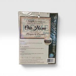 May include: A Dimensions cross-stitch kit with the words "Our Home" in a decorative font. The kit includes cotton thread, a needle, and instructions. The package measures 7 inches by 5 inches.