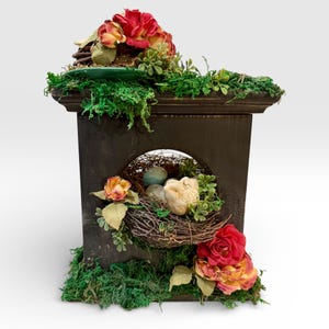 May include: A dark brown decorative birdhouse with a faux nest containing blue eggs and a stone. The birdhouse is adorned with red, yellow, and pink roses, green moss, and greenery. The birdhouse has a decorative archway.