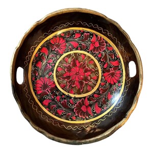May include: A round, wooden serving tray with a dark brown base and a scalloped edge. The tray features a central floral design in red and gold, surrounded by a black background. Two handles are cut into the sides.