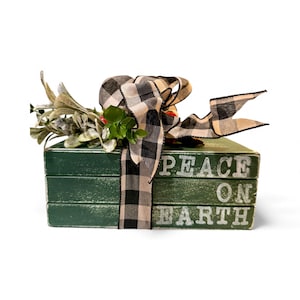 May include: A stack of three green decorative books with the words "PEACE ON EARTH" in white lettering. The books are tied with a black and white checkered ribbon and adorned with faux greenery.