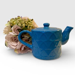 May include: A vibrant blue ceramic teapot with a diamond pattern. The teapot has a curved handle, a spout, and a small knob on the lid. A bouquet of flowers with pink and yellow blooms is next to the teapot.