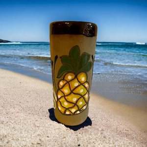 May include: A brown ceramic mug with a painted pineapple design. The mug is on a sandy beach with blue ocean water in the background.