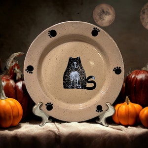 May include: A ceramic plate with a black cat design and paw prints around the edge. The plate is on a stand and surrounded by pumpkins.