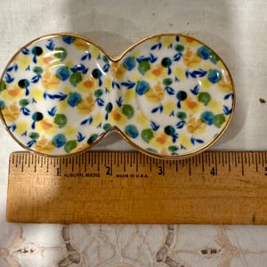 May include: A double-sectioned ceramic dish with a gold rim, decorated with a floral pattern in yellow, blue, and orange. The dish is approximately 5 inches long and is placed next to a wooden ruler.