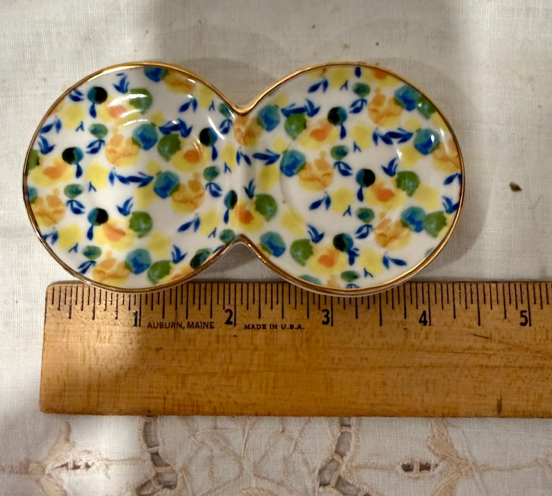 May include: A double-sectioned ceramic dish with a gold rim, decorated with a floral pattern in yellow, blue, and orange. The dish is approximately 5 inches long and is placed next to a wooden ruler.