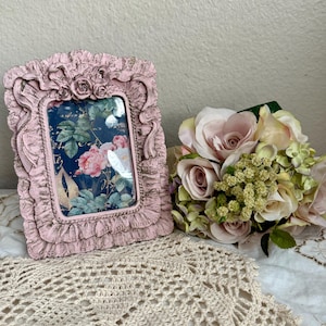 May include: A pink picture frame with a ruffled design and a floral print inside. The frame is sitting on a crocheted doily next to a bouquet of pink roses.