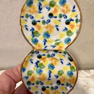 May include: A white ceramic double dish with a gold rim, decorated with yellow, blue, and green floral patterns. The dish has a figure-eight shape, perfect for serving small portions or holding jewelry.