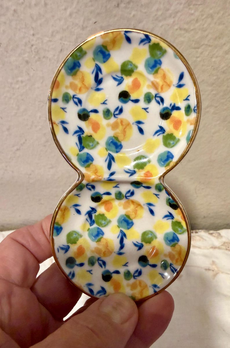 May include: A white ceramic double dish with a gold rim, decorated with yellow, blue, and green floral patterns. The dish has a figure-eight shape, perfect for serving small portions or holding jewelry.