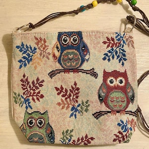 May include: A beige tapestry purse with a brown leather strap and colorful beaded accents. The purse features a repeating pattern of owls and leaves in shades of blue, red, green, and yellow.
