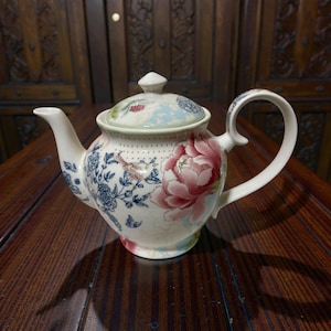 May include: A white ceramic teapot with a lid, spout, and handle. The teapot is decorated with a large pink peony, blue floral patterns, and a bird. The lid features a small knob and a similar floral design. The teapot sits on a wooden surface.