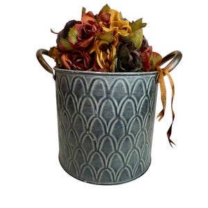 May include: A decorative gray metal bucket with a textured, scalloped pattern, filled with colorful faux flowers in shades of red, orange, and yellow. The bucket has two metal handles and a brown ribbon tied to one side.