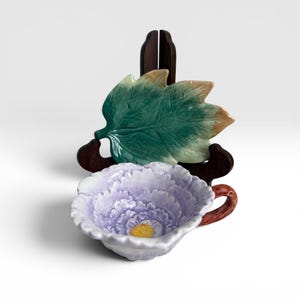 May include: A ceramic teacup and leaf-shaped dish. The teacup is lavender with a yellow center and a brown handle. The leaf-shaped dish is green and brown, resting on a dark wooden stand. The items are likely for serving tea.
