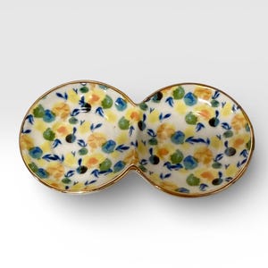 May include: A two-section ceramic serving dish with a floral pattern. The dish has a white base with yellow, blue, and green floral designs. The rim is trimmed with gold. The dish is ideal for serving snacks or dips.