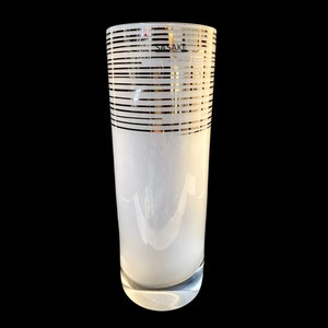 May include: A white frosted glass vase with a gold striped pattern. The vase is tall and cylindrical with a rounded bottom. The word "SASAKI" is printed on the side of the vase.
