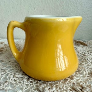 May include: A small, yellow ceramic pitcher with a curved handle and a spout. The pitcher has a glossy finish and a white interior. The pitcher is likely vintage and suitable for serving cream or other liquids.