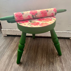 May include: A green wooden stool with three legs and a floral patterned top. A rolling pin with a floral pattern is resting on top of the stool.
