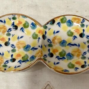 May include: A white ceramic serving dish with two connected oval sections. The dish is decorated with a floral pattern in yellow, orange, blue, and green, with a gold rim. The design features small flowers and leaves.