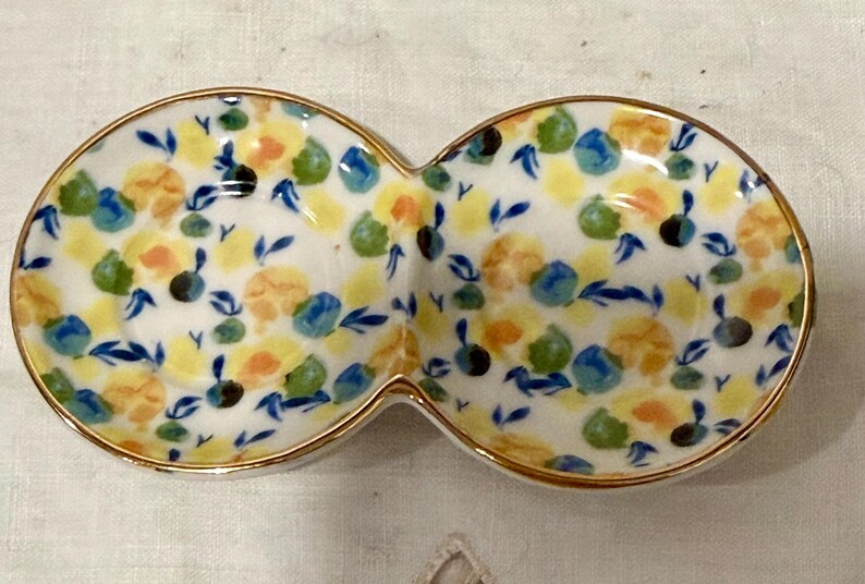 May include: A white ceramic serving dish with two connected oval sections. The dish is decorated with a floral pattern in yellow, orange, blue, and green, with a gold rim. The design features small flowers and leaves.