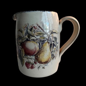 May include: A ceramic pitcher with a cream-colored background and a floral design featuring pears, apples, and strawberries. The pitcher has a blue rim and a handle.