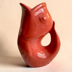 May include: A glossy, coral-colored ceramic pitcher shaped like a stylized fish. The vessel features a wide, open mouth at the top, a smooth, curved body, and a hollow handle formed by the tail section, creating a continuous, fluid silhouette.